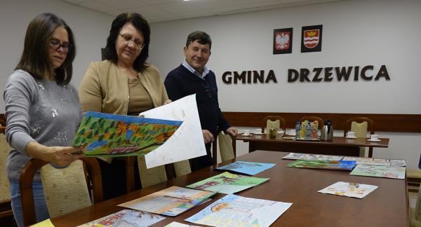 W sali konferencyjnej UM w Drzewicy dwie nauczycielki i Gminny Komendant ZOSP RP oceniają prace plastyczne. Po prawej stronie na ścianie napis Gmina Drzewica, godło Polski i herb Drzewicy