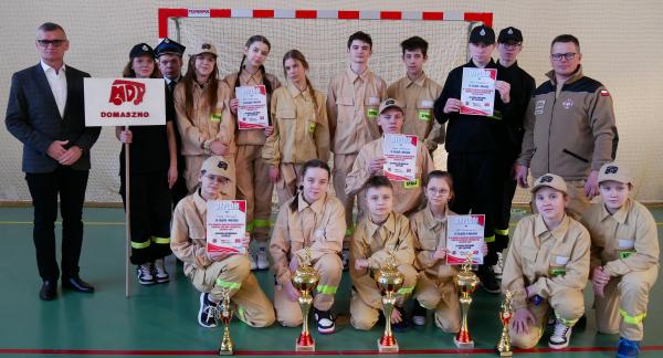 Młodzieżowe Drużyny Pożarnicze z OSP Domaszno wraz z opiekunami prezentują zdobyte trofea. Po lewej stoi Burmistrz Drzewicy.  W tle bramka do piłki ręcznej.