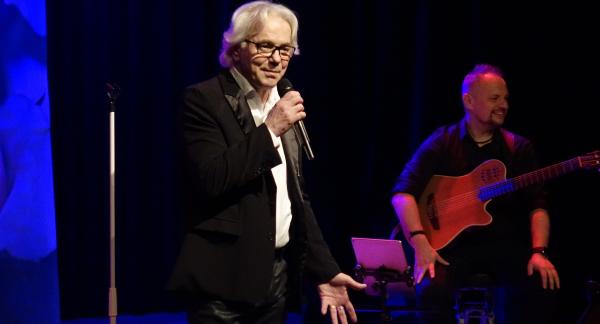 Na scenie z mikrofonem stoi Robert Janowski. Po prawej stronie w tle siedzi gitarzysta.