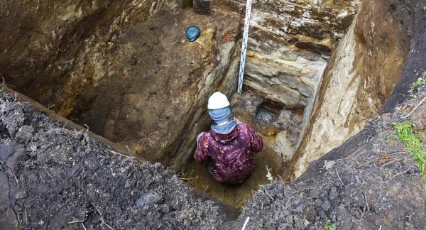 W głębokim wykopie, kucając pracuje archeolog z kaskiem na głowie (widok na jego plecy). Powyżej miarka głębokości.