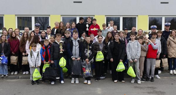 Duża grupa dzieci, młodzieży oraz dorosłych ustawiona w kilku rzędach przed budynkiem o jasnej elewacji z zielonymi elementami. Uczestnicy stoją blisko siebie i pozują do wspólnego zdjęcia. Wśród nich widoczni są uczniowie oraz opiekunowie, a także kilku umundurowanych policjantów