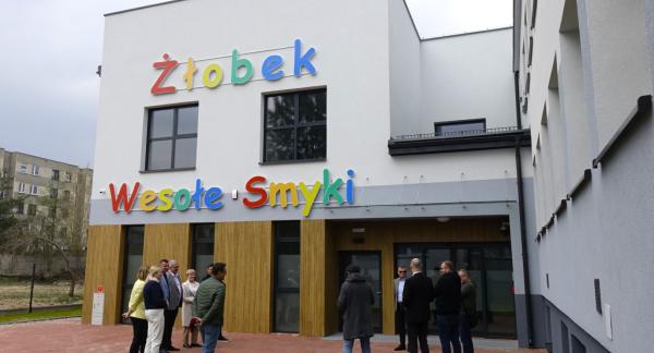 Grupa osób w strojach wizytowych i codziennych stoi przed nowoczesnym, białym budynkiem żłobka „Wesołe Smyki”. Budynek ma kolorowy napis nad wejściem i drewnopodobne panele na parterze.