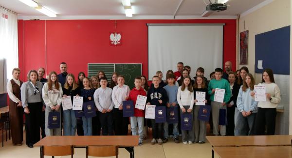 Na zdjęciu grupa uczniów, nauczycieli oraz członków komisji w sali lekcyjnej podczas podsumowania turnieju wiedzy o bezpieczeństwie w ruchu drogowym. Kilka osób trzyma w rękach dyplomy oraz granatowe torby z nadrukiem.
