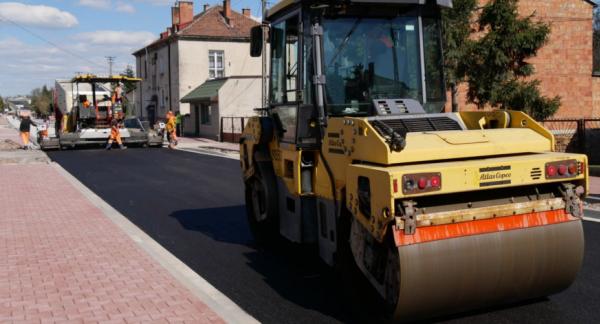 Modernizacja drogi z układaniem nowej nawierzchni asfaltowej przy użyciu specjalistycznych maszyn. Na zdjęciu widoczny walec drogowy oraz pracownicy w odzieży ostrzegawczej podczas prac budowlanych.