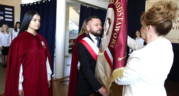 Wnętrze szkoły podczas uroczystości. Mężczyzna z biało-czerwoną szarfą trzyma sztandar szkoły w kolorze czerwonym z haftem. Naprzeciw stoi kobieta w jasnym stroju, poprawiająca lub przekazująca sztandar. Obok  uczennica w czerwonej pelerynie, patrząca w stronę sztandaru. 