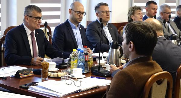 Przy stole z mikrofonami i dokumentami siedzi kilka osób w formalnych strojach – Burmistrz i Zastępca Burmistrza Drzewicy oraz radni. Głos zabiera Burmistrz Drzewicy, pozostali słuchają. Na stole widoczne są napoje i materiały do pracy.