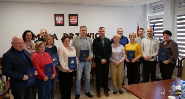 Zdjęcie grupowe przedstawicieli kół gospodyń oraz władz samorządowych w sali konferencyjnej Urzędu Miejskiego w Drzewicy. Uczestnicy trzymają pamiątkowe dyplomy w niebieskich okładkach z herbem gminy.