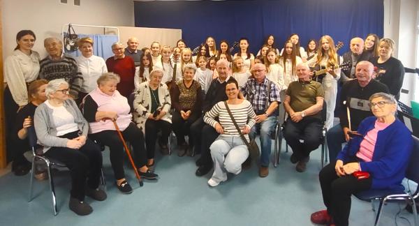 Kilkudzisięcioosobowa grupa seniorów i młodzieży stoi lub siedzi w sali DD Senior+. Młodzi artyści trzymają gitary, wszyscy pozują do wspólnego zdjęcia