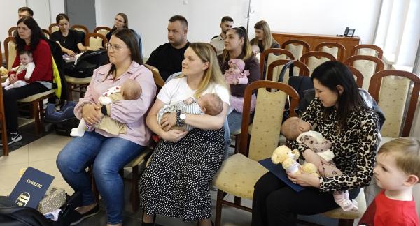 Grupa rodziców z niemowlętami podczas oficjalnego spotkania w Sali USC. W centralnej części siedzą trzy kobiety trzymające na rękach małe dzieci. W tle widoczni są pozostali uczestnicy siedzący na drewnianych krzesłach z jasną tapicerką.