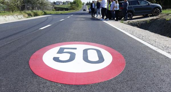 Na pierwszym planie znak poziomy ograniczenia prędkości do 50 km/h namalowany na nowej asfaltowej drodze. W tle po prawej stronie stoi grupa osób przy zaparkowanych samochodach, obserwująca odcinek jezdni. Droga biegnie prosto przez teren zabudowany z domami i zielenią po obu stronach.