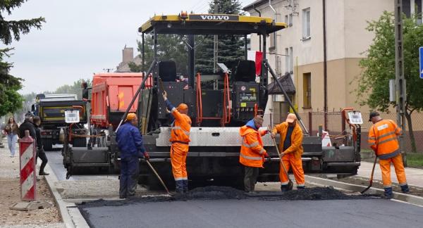 Prace drogowe na ulicy Braci Kobylańskich w terenie zabudowanym. Na środku duża maszyna do układania nawierzchni asfaltowej, za którą kilku pracowników w pomarańczowych strojach ochronnych rozprowadza świeżo ułożony asfalt. W tle samochody ciężarowe, budynki mieszkalne oraz drzewa. Pogoda pochmurna.