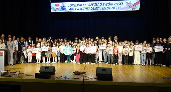  Na scenie sali widowiskowej RCK liczna grupa dzieci, młodzieży, opiekunów i samorządowców stoi w kilku rzędach przed ciemną kurtyną. Niektóre dzieci trzymają symboliczne czeki z nagrodami finansowymi oraz rekwizyty z wcześniejszych występów. Na górze duży biały transparent: „DRZEWICKI PRZEGLĄD TWÓRCZOŚCI ARTYSTYCZNEJ DZIECI I MŁODZIEŻY”. 