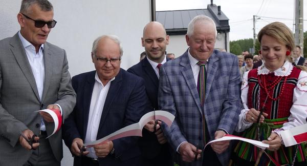  Zbliżenie na moment przecinania biało-czerwonej wstęgi na zewnątrz budynku. Wstęgę przecina pięć osób: Burmistrzowie Drzewicy, Prezes PKP Intercity Remtrak, Sołtys Idzikowic oraz Przewodnicząca KGW Idzikowice w tradycyjnym stroju ludowym.