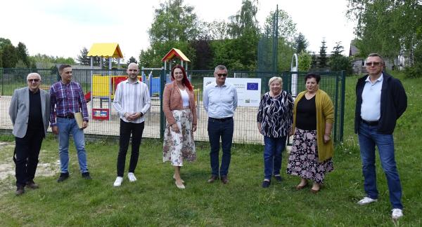 Oficjalne zdjęcie grupowe ośmiu osób stojących na trawie przed ogrodzonym, nowym placem zabaw z tablicą informacyjną w tle.