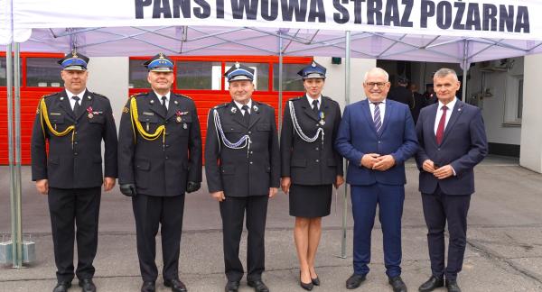 Grupa sześciu osób stojących przed budynkiem pod białym namiotem z napisem „Państwowa Straż Pożarna”. Cztery osoby ubrane są w galowe mundury strażackie, dwie pozostałe w garnitury. W tle znajdują się czerwone bramy garażowe jednostki straży pożarnej. Uczestnicy stoją w jednym rzędzie i pozują do wspólnego zdjęcia.