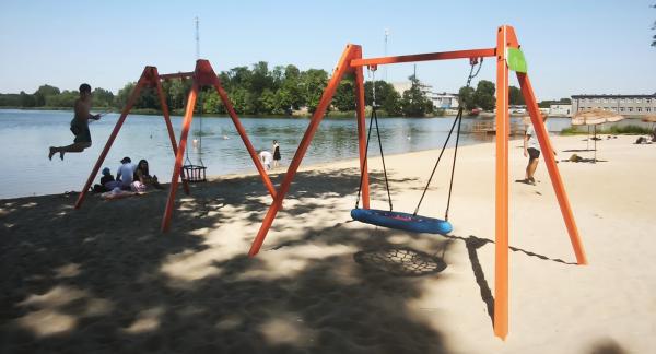 Nowoczesne huśtawki – tradycyjna oraz typu bocianie gniazdo – na pomarańczowych stelażach, ustawione na piaszczystej plaży nad jeziorem. W tle widoczni są kąpiący się i odpoczywający ludzie oraz zielony, przeciwległy brzeg.