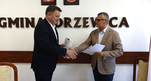 Członek Zarządu Woj. Łódzkiego Artur Bagieński i Burmistrz Drzewicy Janusz Reszelewski stoją w sali konferencyjnej i podają sobie dłonie w uścisku nad stołem, trzymając w rękach dokumenty. W tle biała ściana z dużym napisem „GMINA DRZEWICA”, nad którym wiszą godło Polski oraz herb Drzewicy.