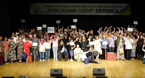 Na scenie w sali widowiskowej RCK kilkadziesiąt osób stoi obok siebie, uśmiechając się i machając rękami. W tłumie widoczne są osoby na wózkach inwalidzkich, osoby w różowych kapeluszach kowbojskich oraz trzymające tabliczki z nazwami różnych placówek (m.in. WTZ). Wysoko duży transparent festiwalu.
