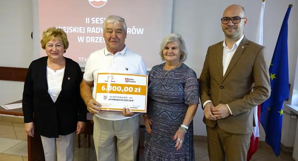 Pozowane zdjęcie czterech osób stojących obok siebie w sali konferencyjnej. Drugi od lewej mężczyzna trzyma duży, symboliczny czek na kwotę 6000,00 zł na realizację projektu „WYPRAWA PO ZDROWIE”. Po prawej stronie stoi mężczyzna w jasnobrązowym garniturze i okularach. W tle widoczny jest ekran projekcyjny z napisem „II SESJA MIEJSKIEJ RADY SENIORÓW W DRZEWICY” oraz flagi państwowe.