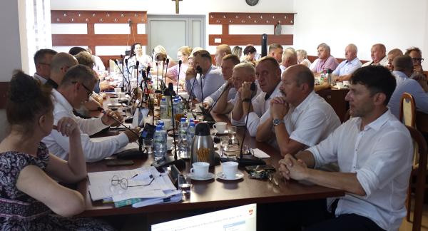 Szeroka panorama sali konferencyjnej Urzędu Miejskiego w Drzewicy. Przy długim stole siedzi kilkunastu mężczyzn i kobiet ubranych oficjalnie. Uczestnicy sesji, w tym Burmistrz Janusz Reszelewski, skupieni są na dokumentach i ekranie, na którym widoczny jest porządek obrad XXV sesji Rady Miejskiej. Na stole znajdują się mikrofony, butelki z wodą i filiżanki.