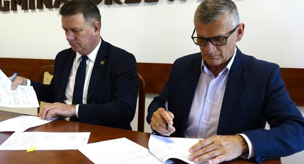 Członek Zarządu Woj. Łódzkiego Artur Bagieński i Burmistrz Drzewicy Janusz Reszelewski w garniturach siedzą obok siebie przy drewnianym konferencyjnym stole. Obaj podpisują leżące przed nimi dokumenty. Na ścianie w tle widoczny duży, brązowy, wypukły napis „GMINA DRZEWICA”.