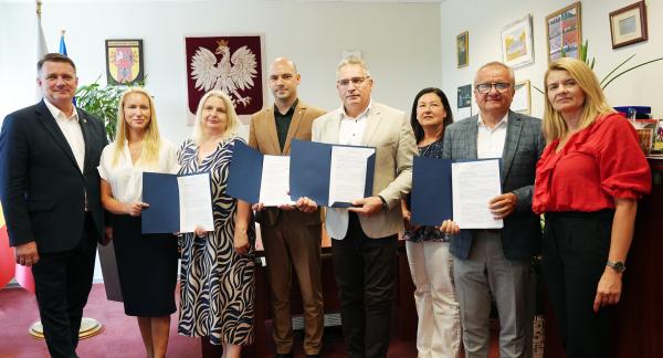 Grupa ośmiu osób w gabinecie pozuje do wspólnego zdjęcia. Cztery osoby prezentują otwarte niebieskie teczki z podpisanymi dokumentami. W środku Zastępca Burmistrza Drzewicy Dominik Niemirski.
