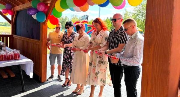 roczyste przecięcie czerwonej wstęgi pod drewnianą wiatą w Domasznie, przyozdobioną kolorowymi balonami podczas Pikniku Rodzinnego.