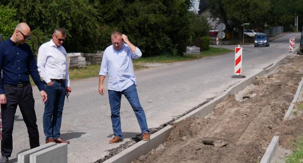 Trzech mężczyzn w strojach wizytowych ogląda plac budowy drogi ze świeżo ułożonymi krawężnikami na pierwszym planie oraz pracującą w oddali koparką