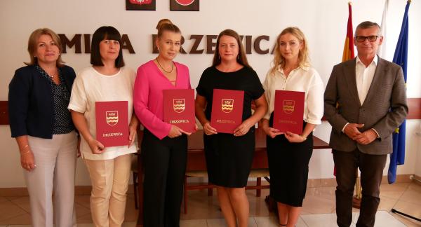 Zdjęcie grupowe sześciu osób stojących w sali konferencyjnej Urzędu Miejskiego w Drzewicy. Cztery nauczycielki w środku trzymają czerwone teczki z herbem Drzewicy. Po lewej Dyrektorka ZEAS, po prawej Burmistrz Drzewicy. Na ścianie Godło Polski i herb Drzewicy oraz przysłonięty napis Gmina Drzewica. Za Burmistrzem flagi.