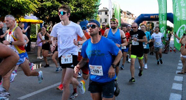 Grupa biegaczy w trakcie wyścigu ulicznego Drzewica Summer Run. Na pierwszym planie mężczyzna w niebieskiej koszulce z numerem 109 i okularach przeciwsłonecznych, obok biegacz w białej koszulce.