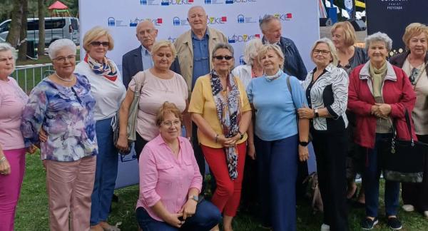 Grupa kilkunastu seniorów reprezentujących Gminę Drzewica pozuje do pamiątkowej fotografii w plenerze na tle promocyjnej ścianki Województwa Łódzkiego podczas Wojewódzkich Obchodów Dnia Seniora