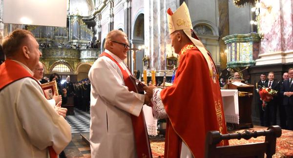 Uroczystość kościelna we wnętrzu świątyni w Studziannie. Biskup Marek Solarczyk w czerwonej szacie liturgicznej i mitrze wręcza akt nominacji ks. Grzegorzowi Włodarczykowi w białej albie i czerwonej stule. Z lewej strony przygląda się inny duchowny. W tle zabytkowe organy, nawy kościoła oraz zgromadzeni wierni i strażacy. 