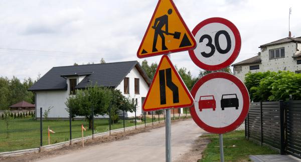 Znaki drogowe ostrzegające o robotach drogowych, zwężeniu jezdni, ograniczeniu prędkości do 30 km/h oraz zakazie wyprzedzania, ustawione przy remontowanej drodze w sąsiedztwie domów jednorodzinnych.