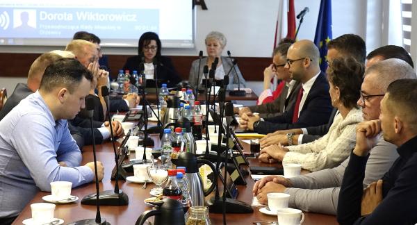 Sala konferencyjna UM w Drzewicy, w której odbywa się posiedzenie. Przy długim stole, na którym ustawiono mikrofony, filiżanki, butelki z napojami oraz dzbanki, siedzą po obu stronach radni i uczestnicy obrad. Na dalszym planie, na przedzie sali, zasiadają członkowie Prezydium Rady Miejskiej. Za nimi ekran projekcyjny oraz flagi.   