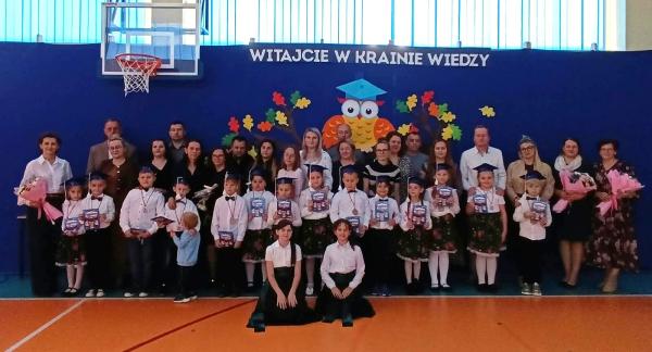 Grupowo pozujące dzieci w galowych strojach, nauczyciele i rodzice podczas uroczystości szkolnej w sali gimnastycznej SP w Brzustowcu. W tle niebieska ściana z napisem „WITAJCIE W KRAINIE WIEDZY” oraz dekoracją z sową i jesiennymi liśćmi.