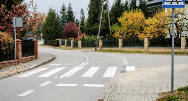 Przejście dla pieszych na łuku asfaltowej drogi z pylonami i znakiem D-6 w otoczeniu jesiennej zieleni i domów jednorodzinnych.
