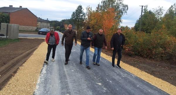 Pięciu mężczyzn idących nowo wybudowaną drogą asfaltową w Brzustowcu-Smugach w otoczeniu zabudowań i jesiennej zieleni. 