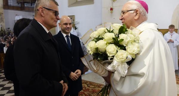 Arcybiskup Wacław Depo w białych szatach liturgicznych i różowej piusce przyjmuje duży bukiet białych róż wewnątrz kościoła. Naprzeciw niego stoi Burmistrz Drzewicy w ciemnym płaszczu i okularach, a obok niego Wiceburmistrz Drzewicy w garniturze.
