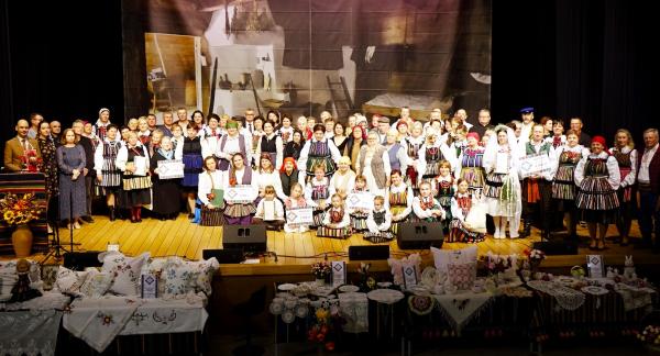Zdjęcie grupowe uczestników Rewii na scenie sali widowiskowej RCK. W centrum osoby w tradycyjnych strojach ludowych, dzieci oraz goście. Na pierwszym planie stoły nakryte haftowanymi obrusami z wyrobami rękodzieła i poduszkami dekoracyjnymi. W tle dekoracja sceniczna z motywem dawnej chaty.