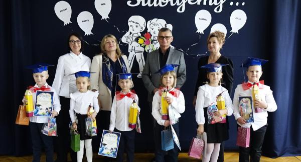  Sześcioro dzieci w biretach i strojach galowych pozujących do wspólnego zdjęcia z dyrekcją, kadrą pedagogiczną i gośćmi na tle dekoracji z napisem „Uroczyście Ślubujemy...”.