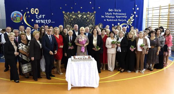 Pozowane zdjęcie grupowe kilkudziesięciorga uczestników jubileuszu 60-lecia SP w Radzicach Dużych. Przed grupą stoi stolik z uroczystym tortem, a w tle znajduje się niebieska ścianka dekoracyjna. 