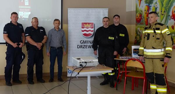 Grupa sześciu mężczyzn (policjanci, strażacy, cywil) stoi w rzędzie w sali prelekcyjnej między dwoma banerami reklamowymi a ekranem projekcyjnym.