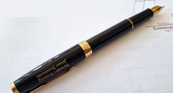  Eleganckie, czarne wieczne pióro ze złotą stalówką i złotymi wykończeniami, leżące na białym dokumencie papierowym. Na korpusie pióra znajduje się grawerowany napis w kolorze złotym: „Burmistrz Drzewicy Janusz Reszelewski”. W tle widoczny jest fragment podpisu i pieczątki na dokumencie.